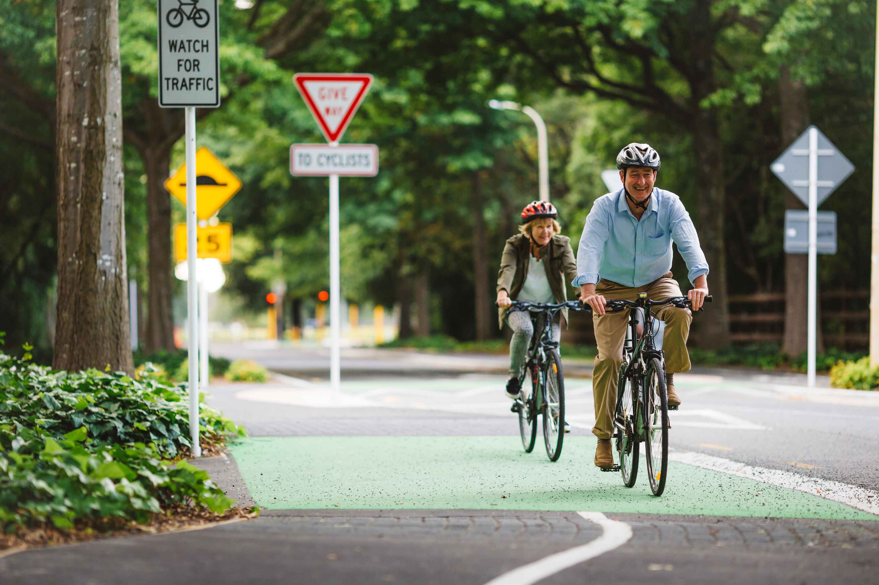 Older man smiling wearing a helmet and cycling towards the viewer. Followed close behind by a woman also on a bike wearing a helmet.